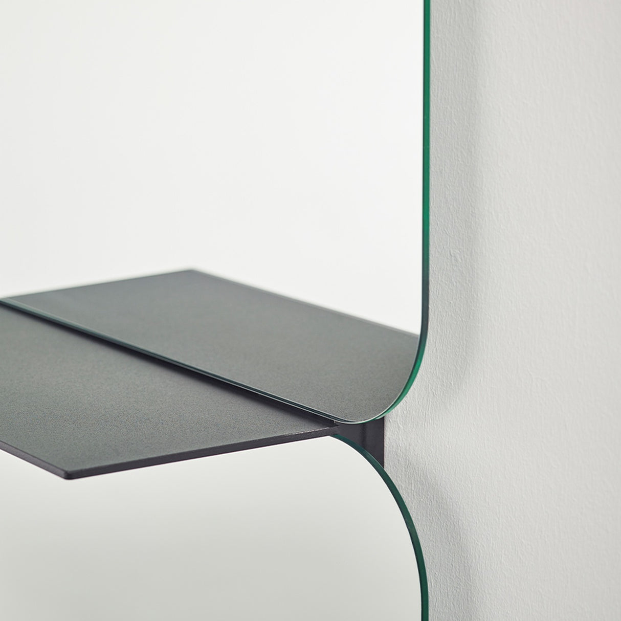 Close-up of the Taille full length hall mirror, showing two frameless rectangular glass panes with rounded corners meeting at a folded black metal shelf, against a plain white wall.