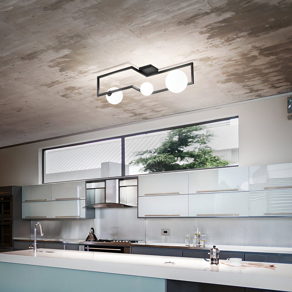 A modern kitchen is equipped with sleek, minimalist cabinetry adorned with metallic finishes and a central island. To illuminate the space, it features the contemporary Dazzle 3 Light Flush in Black as the ceiling light fixture. Large windows invite natural light and offer expansive views of the greenery outside.