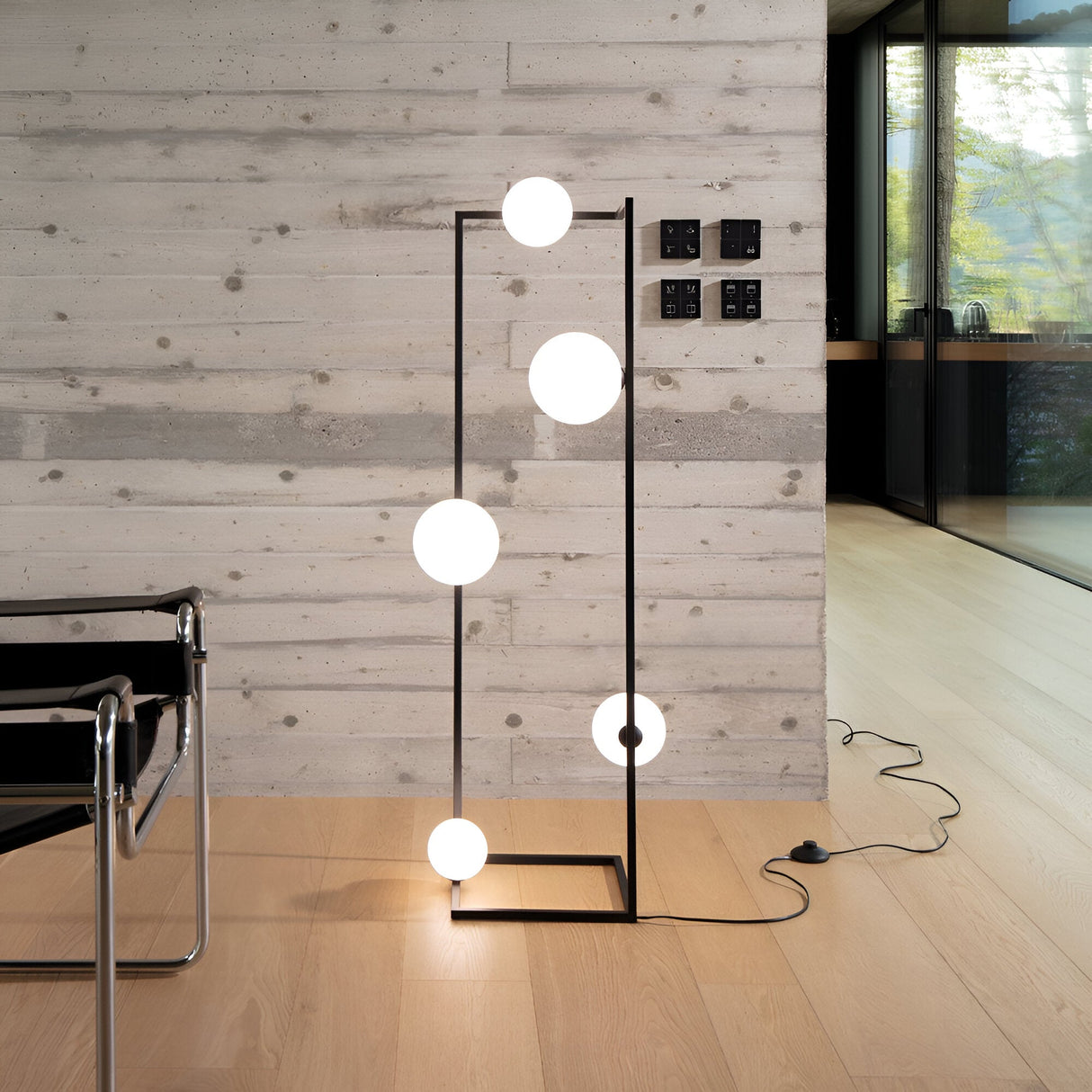 The Dazzle Floor Lamp - Black/White, showcasing a geometric design, incorporates six glowing orbs within a sleek black metal frame. It emits an ambient light while standing on a light wooden floor against a textured gray concrete wall, with part of a black chair visible to the left.