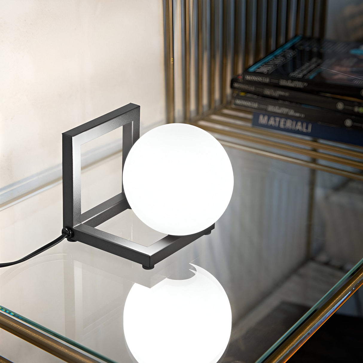 The Dazzle Table Lamp - Black, featuring a spherical white light and a sleek black finish, rests on a glass shelf. Its minimalist metal frame embodies modern elegance, while stacked books with dark covers nearby accentuate the contemporary interior design.