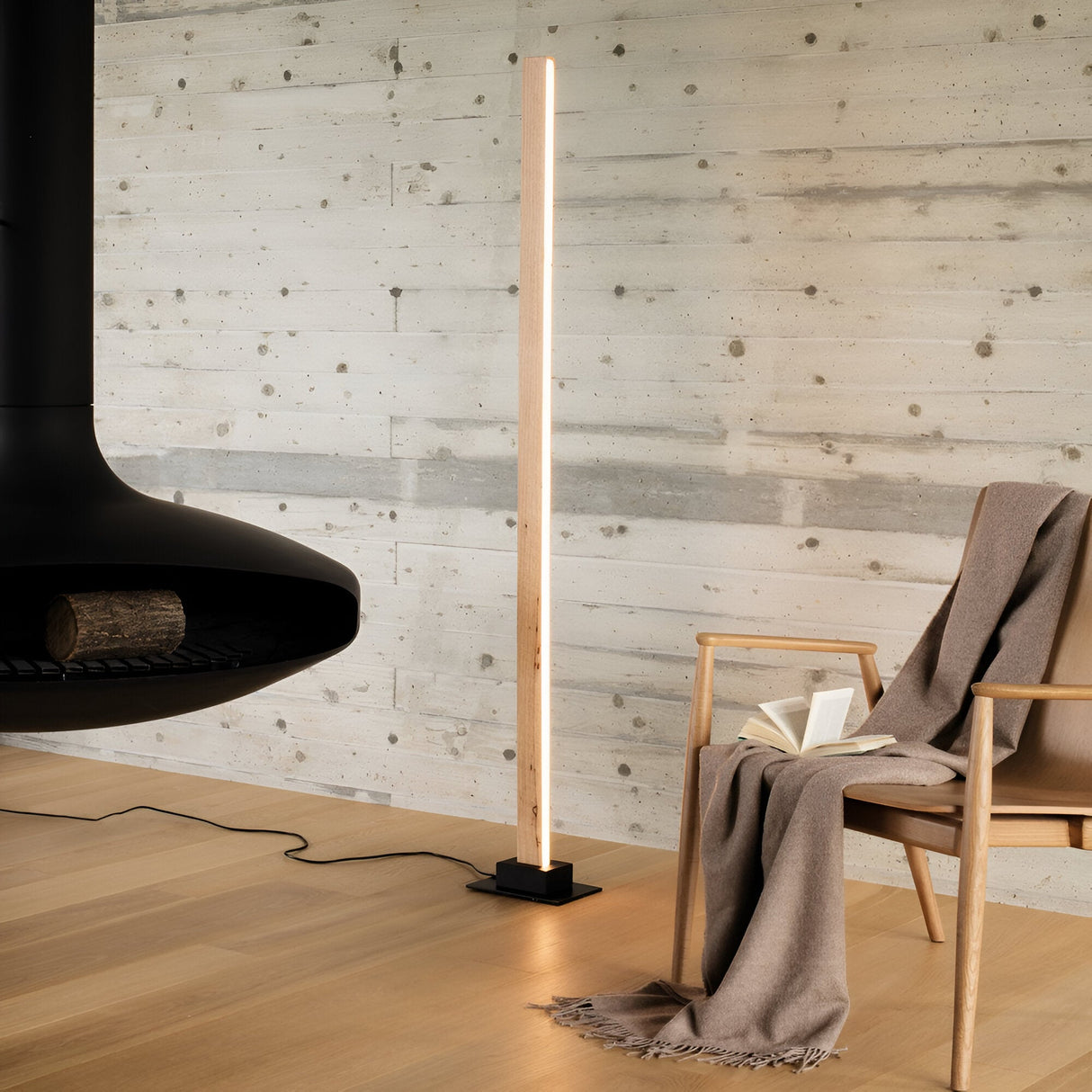 In a modern living room with a minimalist aesthetic, the Erin Wood LED Floor Lamp 26W 3000K in light brown stands elegantly on a natural wood floor beside a cozy chair draped with a blanket and an open book. A suspended black fireplace with a piece of log rests against a textured concrete wall, featuring energy-efficient lighting.
