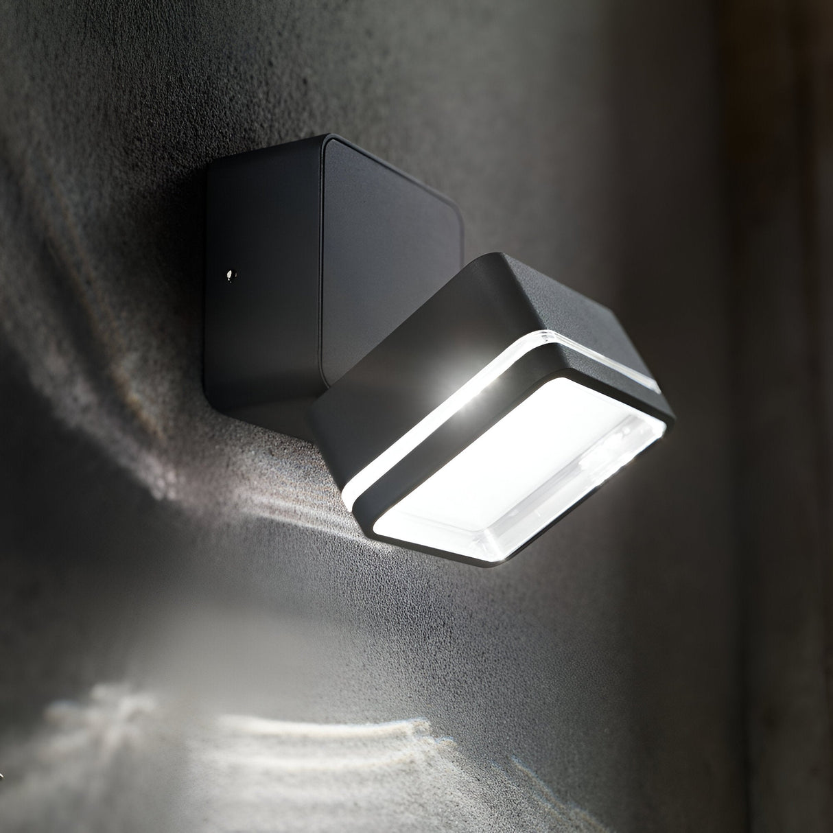The Arcane Square Outdoor LED Single Spotlight 7W 4000K in grey is illuminated, casting an energy-efficient glow onto a textured wall. This weather-resistant light showcases a sleek square design, with its illumination enhancing the surface below.