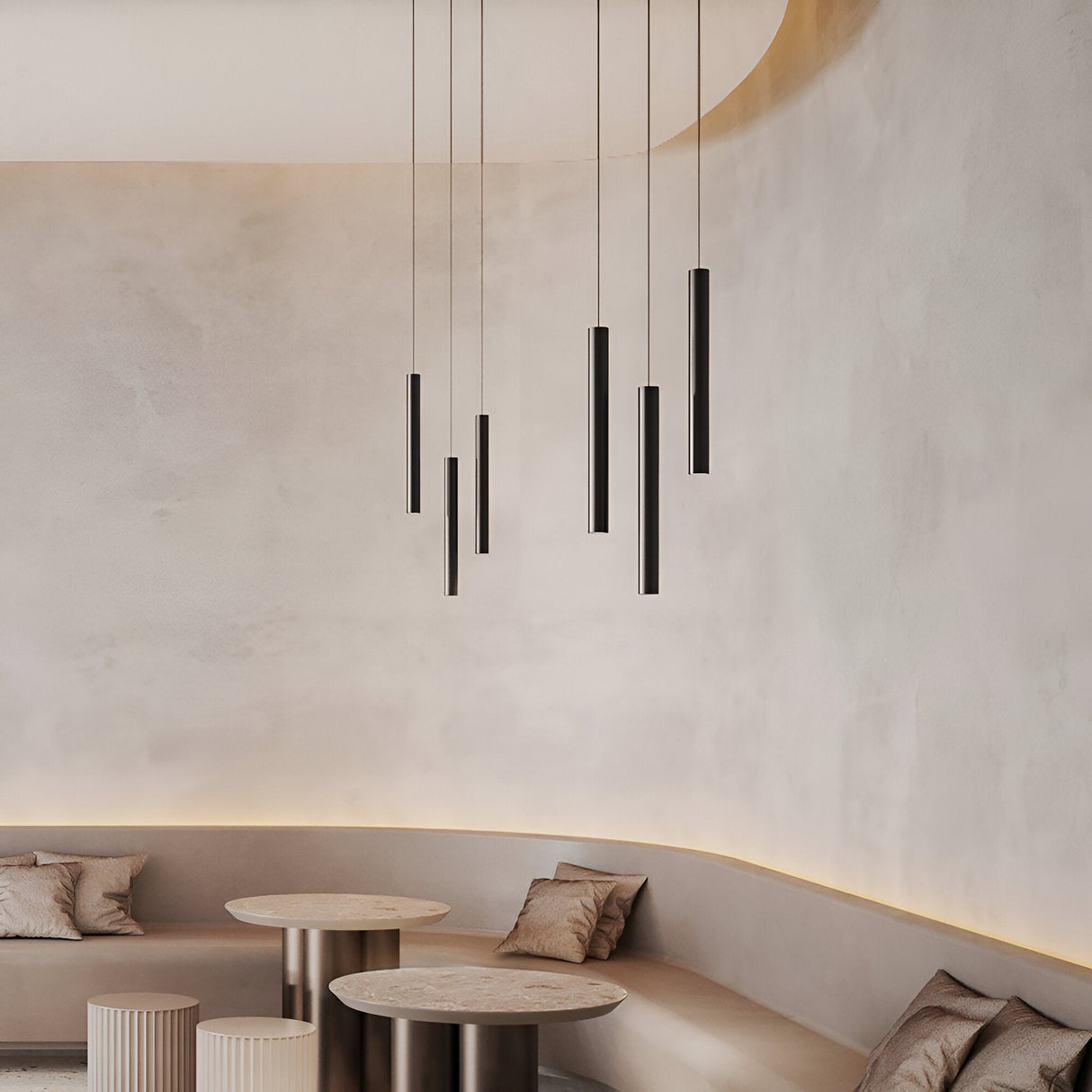 A minimalist room with a soft beige color scheme features rounded seating areas adorned with gray cushions. Hanging from the ceiling are several sleek Pure Dimmable LED Magnetic Track Pendant lights in black, providing a warm 3000K glow. In front of the benches are two small round tables, complementing the overall aesthetic.