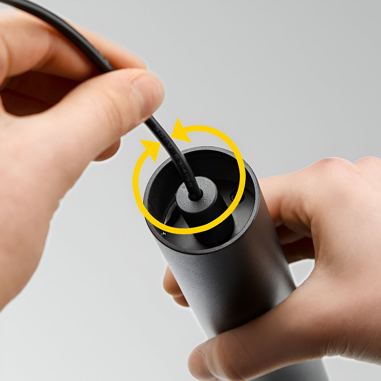 Two hands are shown inserting a black cable into the sleek, cylindrical Pure Dimmable LED Magnetic Track Pendant 12W 3000K device, with yellow arrows indicating a twisting motion. The background is gray and plain, emphasizing the modern lighting design.