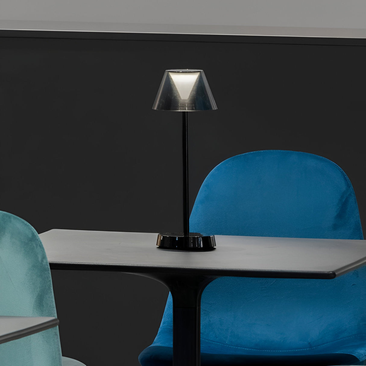 The Solitus LED Outdoor Table Lamp 2.8W 3000K in black, with its modern design, sits at the center of a table surrounded by plush blue chairs. Its IP54 rating ensures durability against the elements, making it ideal for both indoor and outdoor use, while the rechargeable battery adds to its versatility.