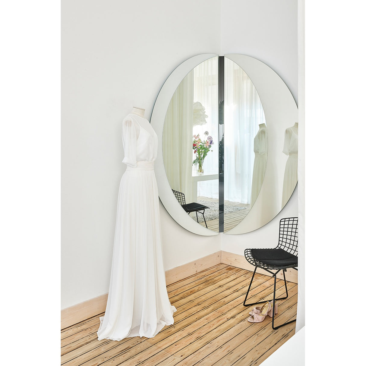 Luna Black Two Piece Corner Wall Mirror 150cm, a round frameless mirror split across a room corner with a black lacquered glass strip, shown beside a dressed mannequin and wire chair.