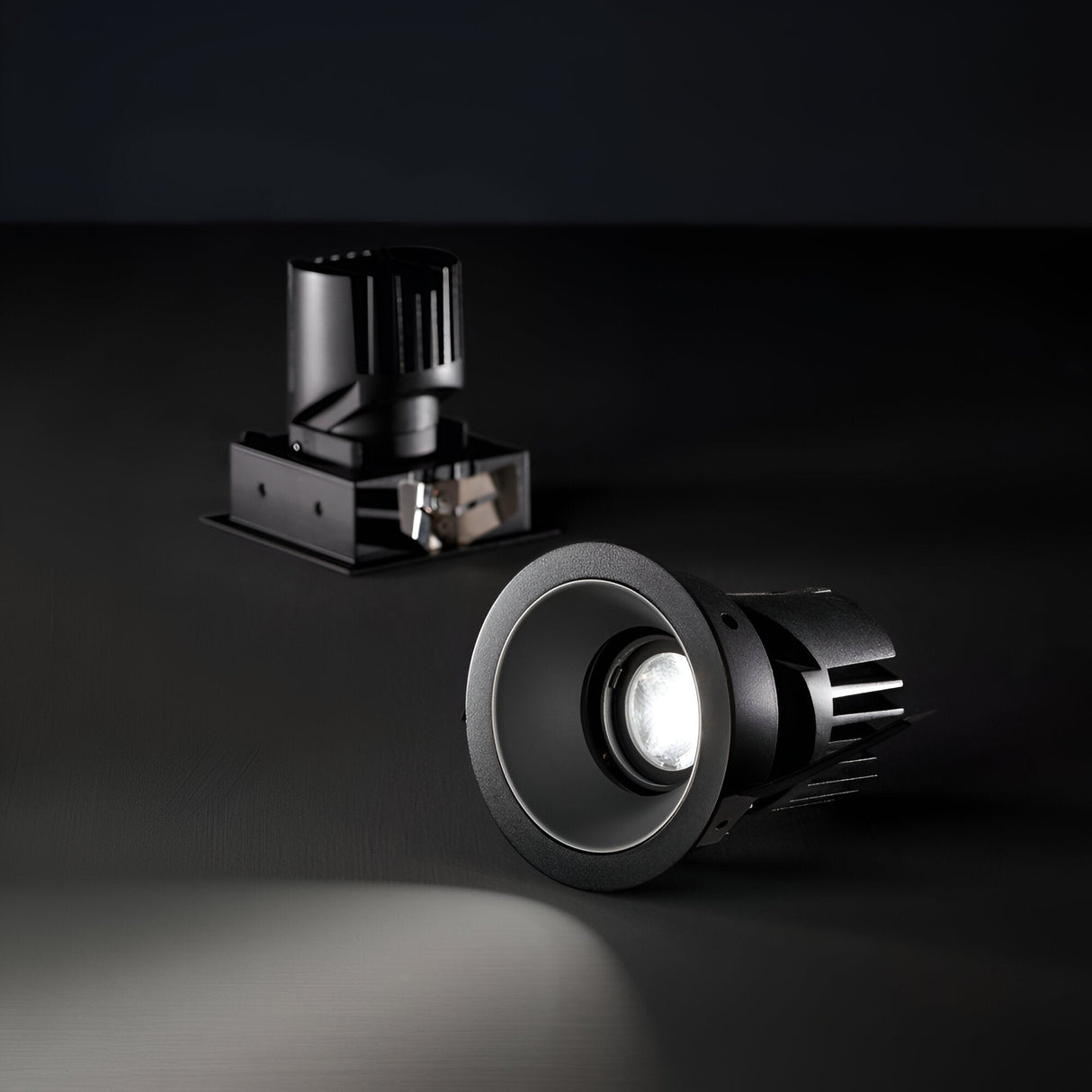 Two black recessed LED ceiling lights sit on a dark surface. The Voltis Round Frame 107mm - Black is pointed towards the camera, casting light on its surroundings. The other light is positioned slightly behind and remains unlit, highlighting its cylindrical housing and mount details—an excellent example of modular lighting design.