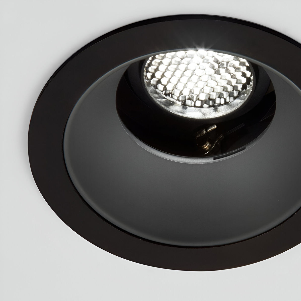 A close-up view of a black recessed ceiling light highlights its Voltis Round Frame 107mm - Black with an illuminated LED bulb. This modular lighting solution is set into a white ceiling, casting a bright and even glow.