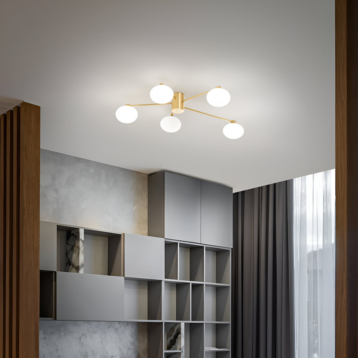 A modern room showcases the Enigma 5 Light Semi Flush fixture in brass, measuring 90cm, along with gray shelves featuring a geometric design, wood paneling, and dark curtains. Ample natural light spills through a sheer window, highlighting the contemporary decor. The dimmable lighting adds versatility to the ambiance.