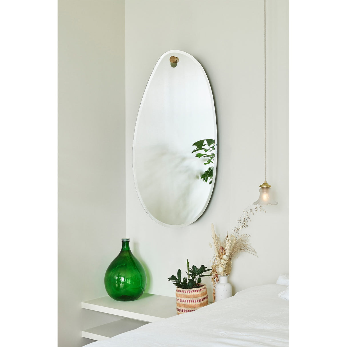 Globo Lago Oak Peg Hung Wall Mirror, a frameless organic-shaped mirror with a bevelled edge, hung from a solid oak peg on a light bedroom wall above a shelf with decor.