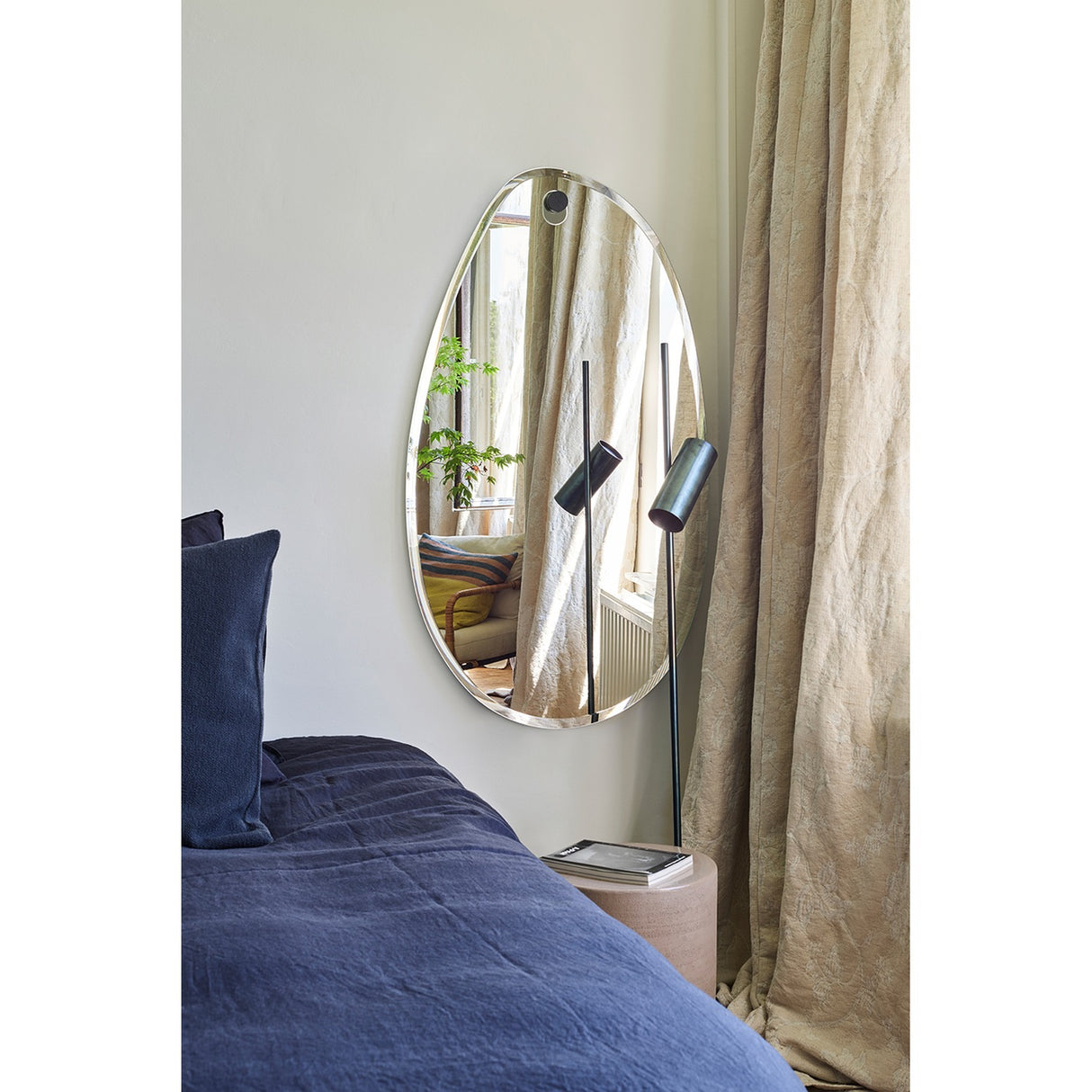 Globo Lago Oak Peg Hung Wall Mirror, a frameless organic-shaped mirror with bevelled edge, shown above a bed reflecting curtains and a floor lamp in a bedroom setting.