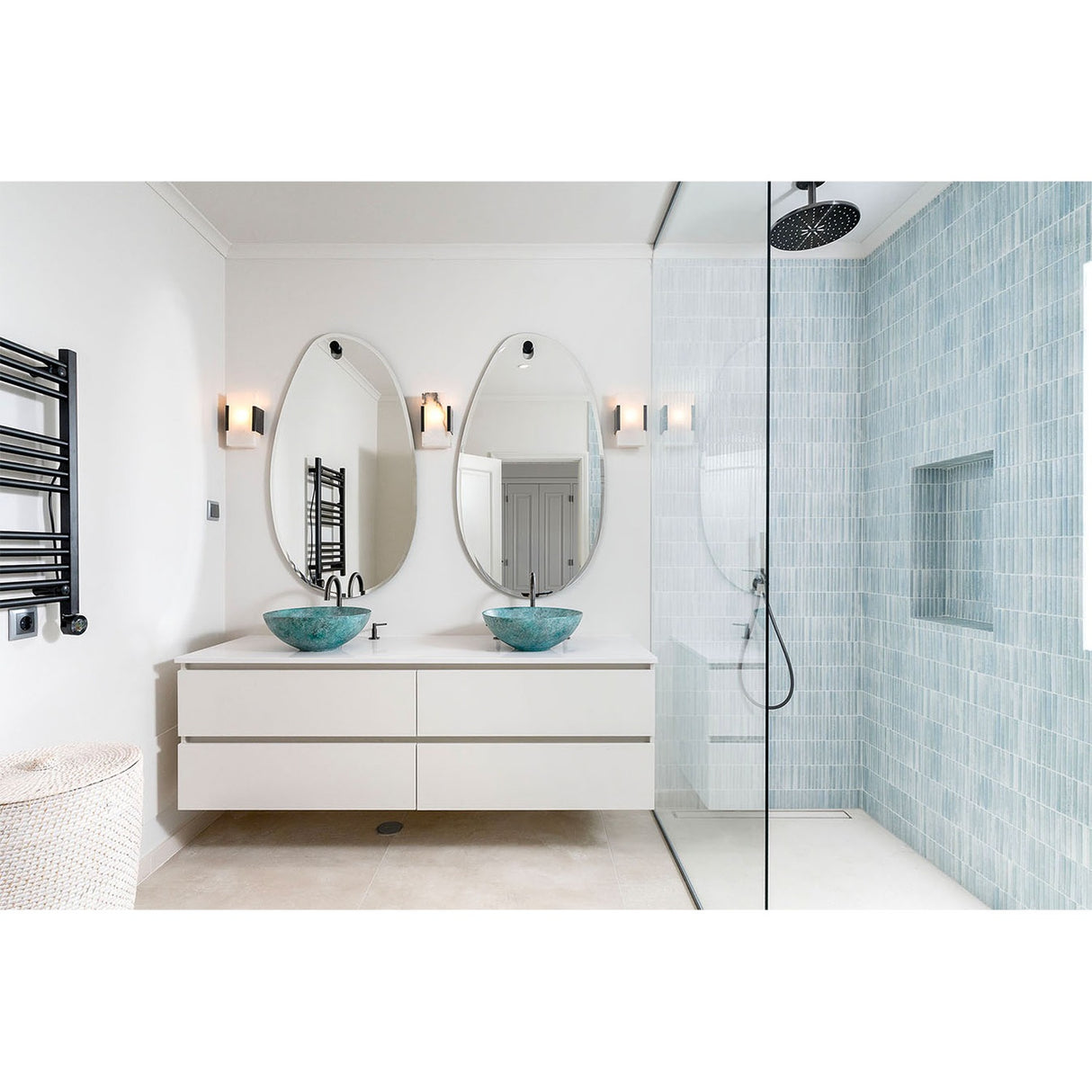 Two oval frameless wall mirrors above a bathroom vanity with teal vessel sinks, hung from black wall fittings against white walls and blue tiled shower.
