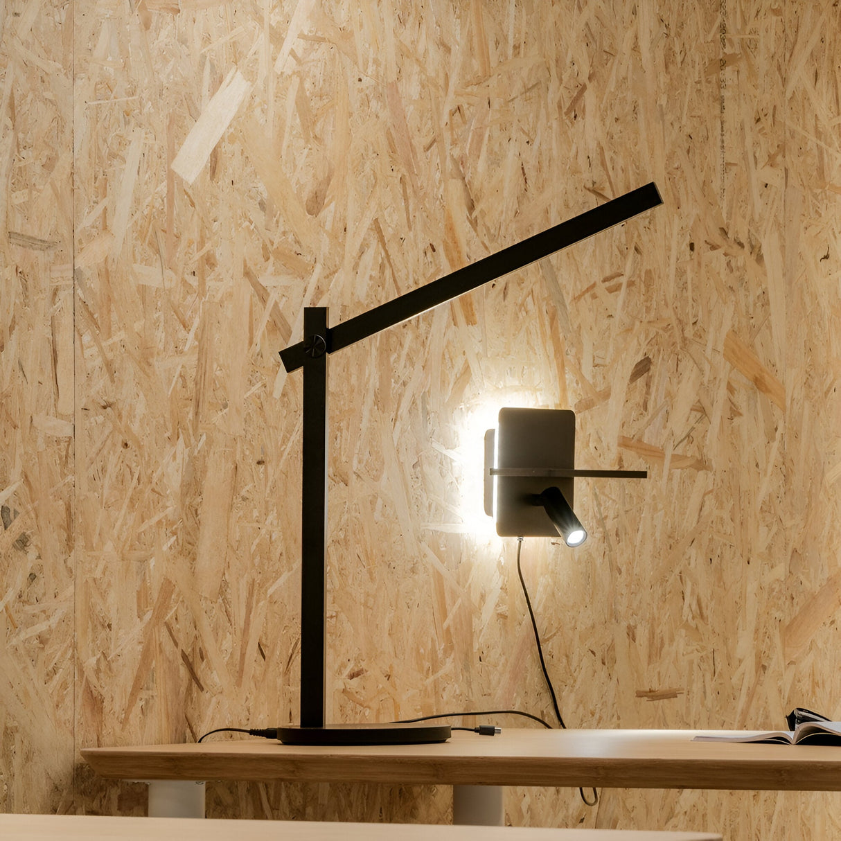 The Arcanta LED Table Lamp in black, featuring a modern minimalist design with an adjustable arm and diffuser, casts light against a textured wooden wall. An open book rests on a light-colored wooden desk beneath the sleek form of the lamp.