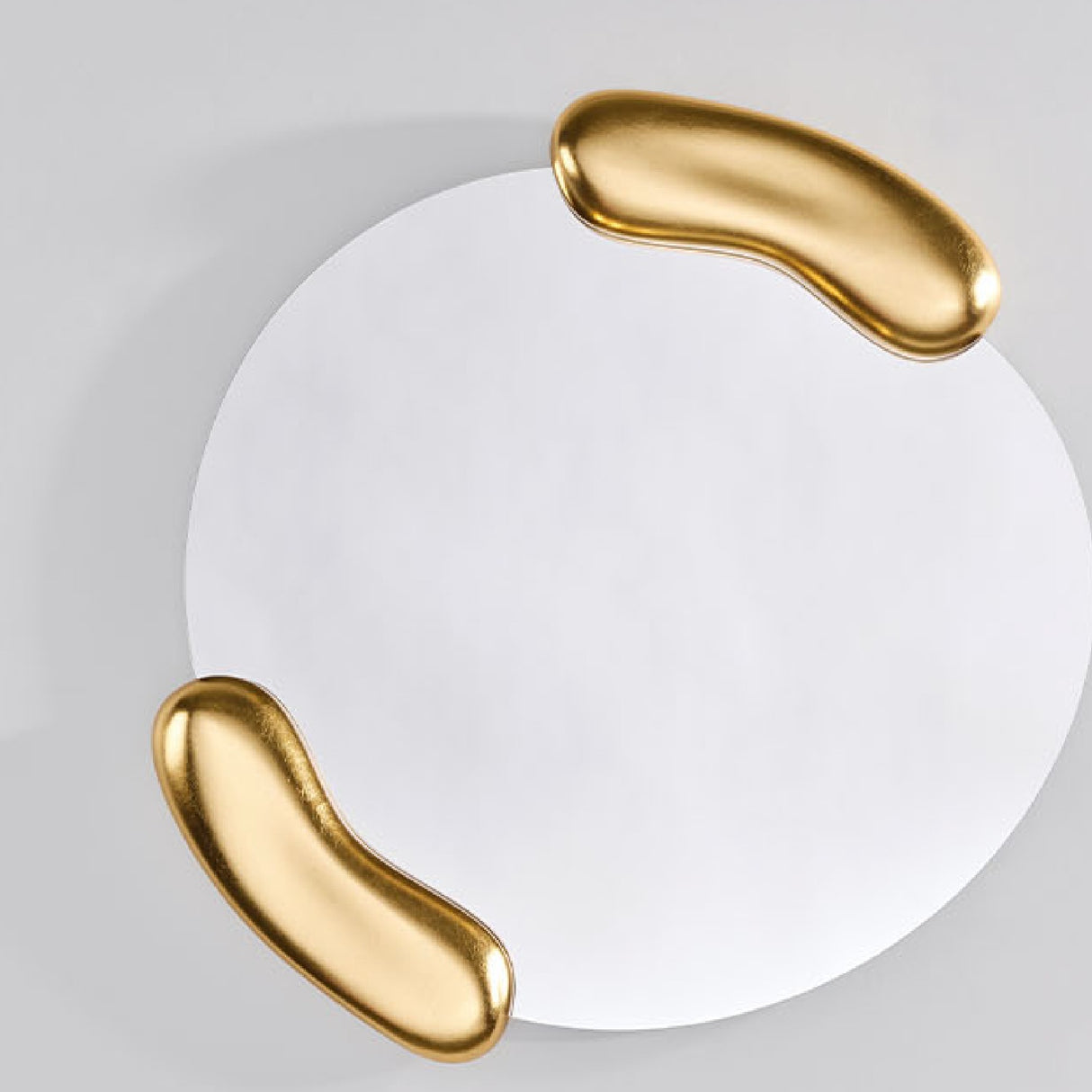 Calco Gold Round wall mirror, 73cm, with an asymmetric glass shape framed by two sculpted gold leaf forms holding it at opposite sides, shown against a plain light grey background.
