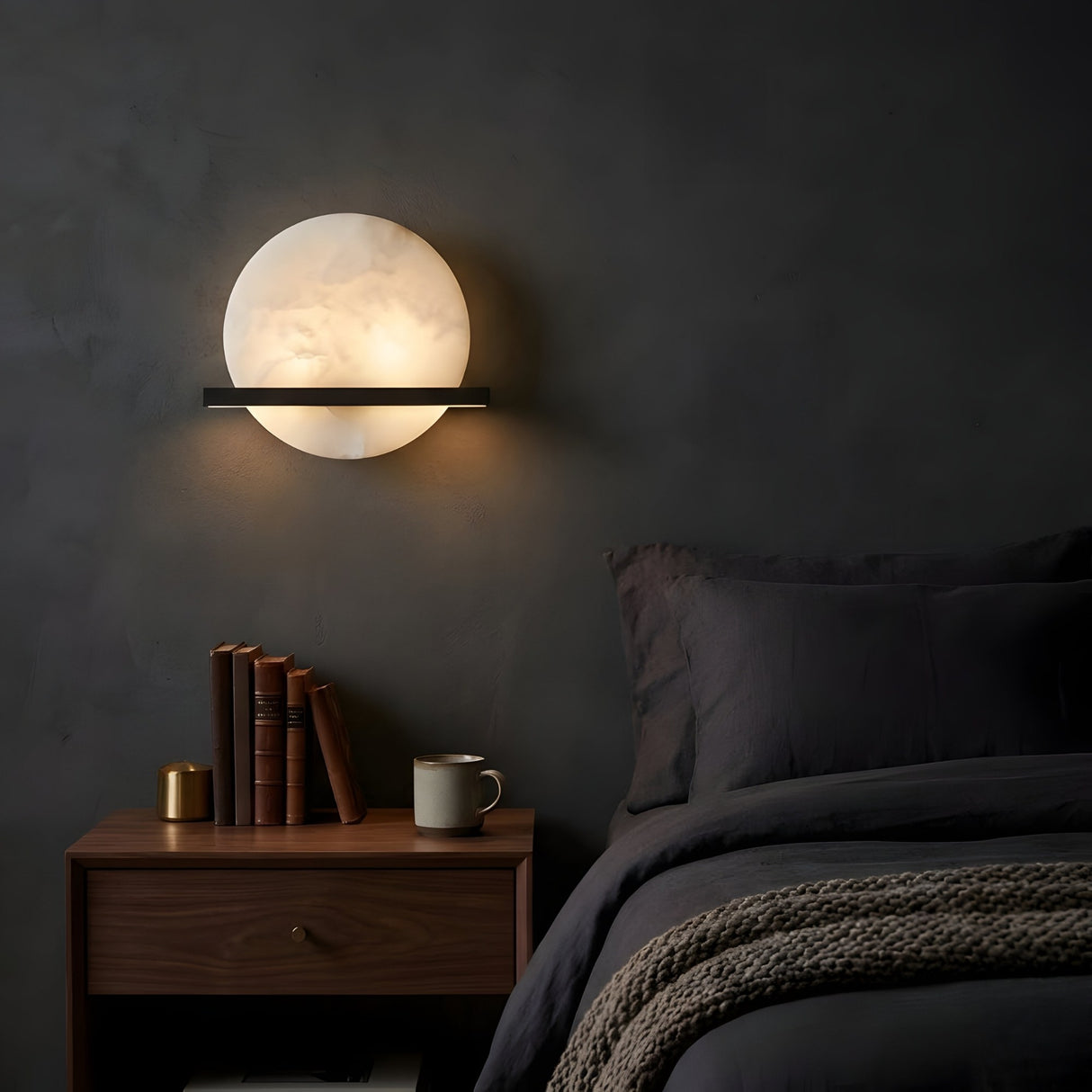 A modern bedroom with dark walls features the Caldris LED Circular Alabaster Wall Light in Matte Black & Soft White above a wooden nightstand, books, a brass object, mug, and a neatly made bed with dark bedding and a knitted blanket.