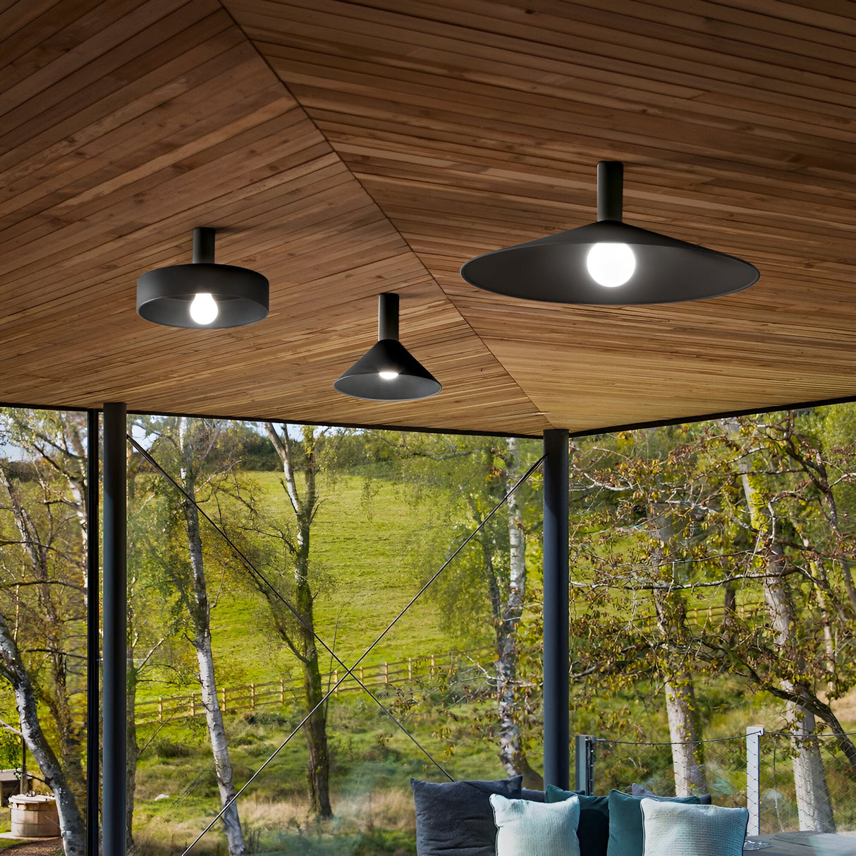 Hanging from the wooden paneled roof on a patio, three Arion Ceiling Lights in black, complete with dimmable features, illuminate the space. Large windows frame a lush outdoor view of trees and grass, while a cozy corner adorned with cushions sits snugly beneath the lights.