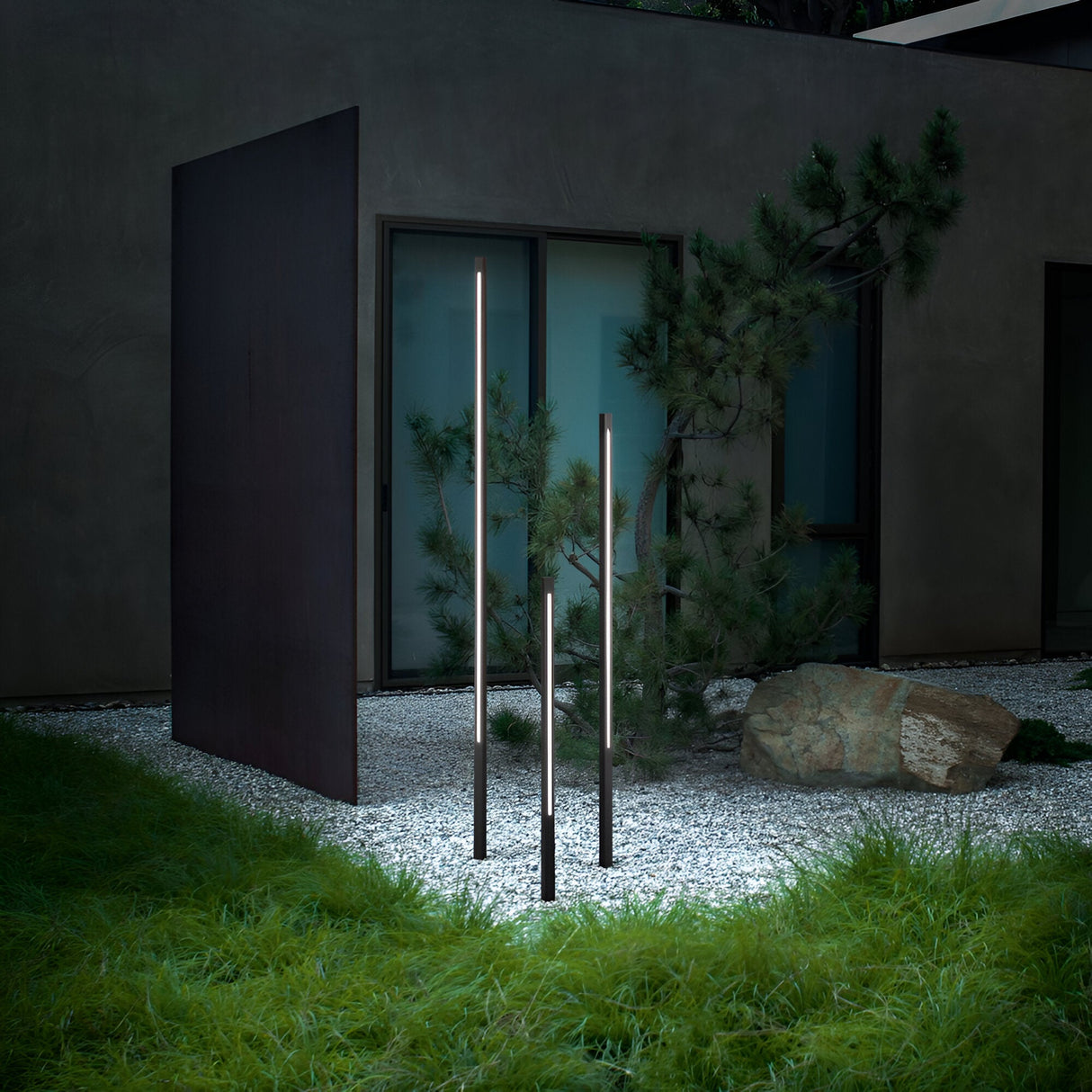 A modern garden with a contemporary design features three Arcera Outdoor LED Bollard Lights in black, standing at 120cm tall. These sleek, energy-efficient vertical light poles are elegantly placed among gravel and a small tree, adjacent to a dark-paneled wall. Additionally, a large rock enhances the serene and natural ambiance of the space.