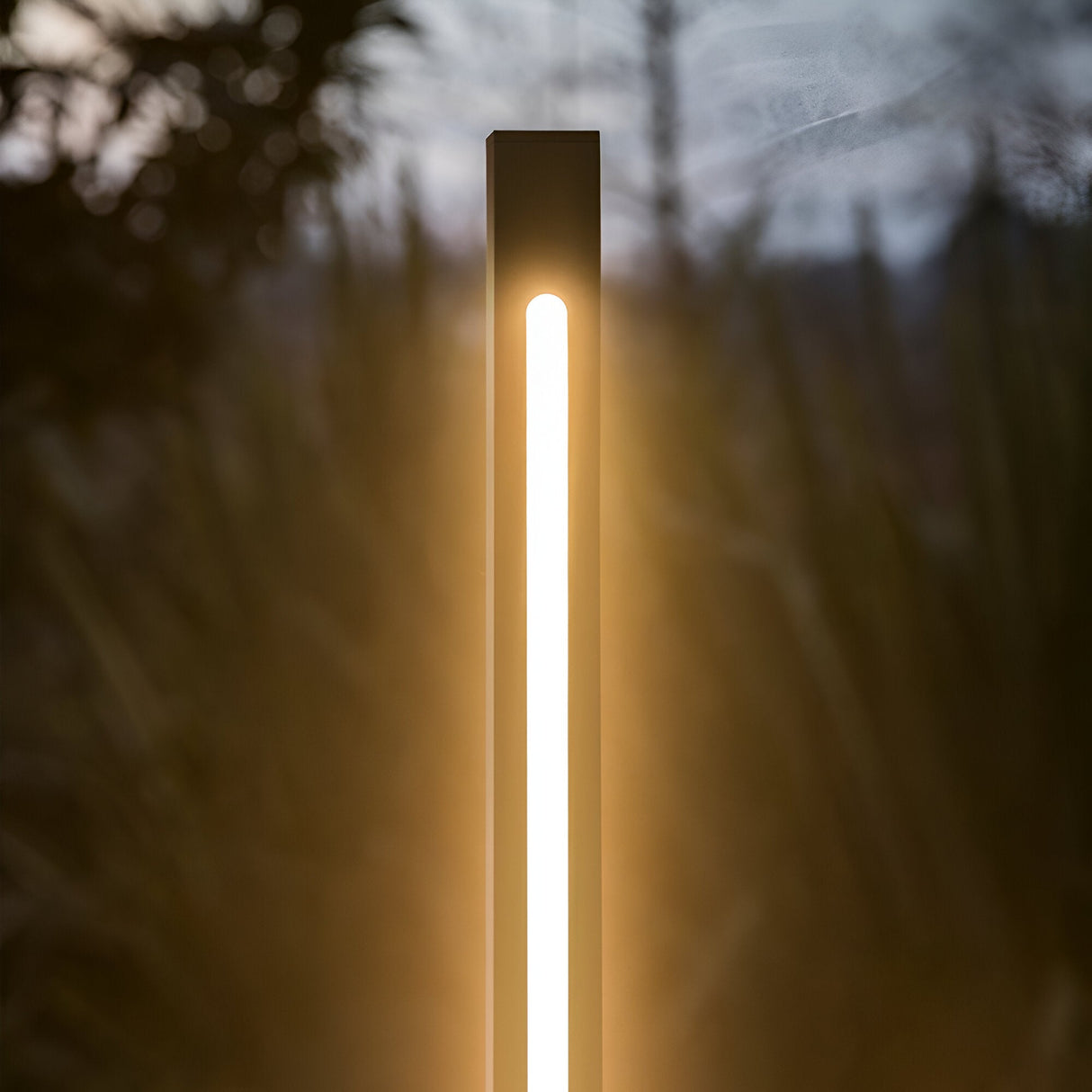 Against a backdrop of dark foliage, the sleek Arcera Outdoor LED Bollard Light in black stands tall at 160cm, emitting a soft, warm white glow that enhances the ambient evening atmosphere.