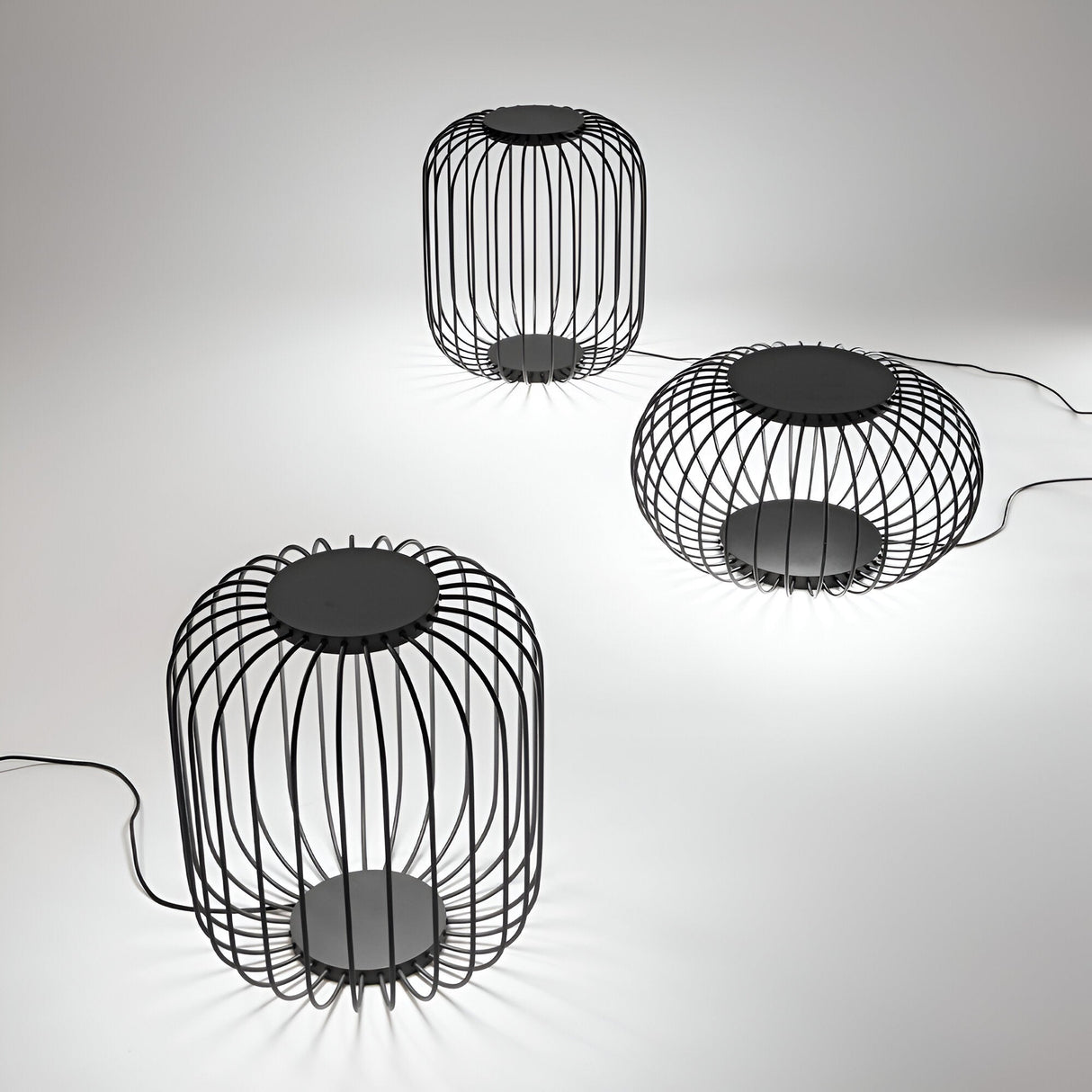 Three Luxtra Outdoor LED Floor Lamps, each boasting a modern black wireframe design and sleek finish, are arranged on a white surface. These lamps have circular bases and tops, casting a soft glow that complements the minimalist setting. Visible cords connect them to a power source for optimal energy efficiency.
