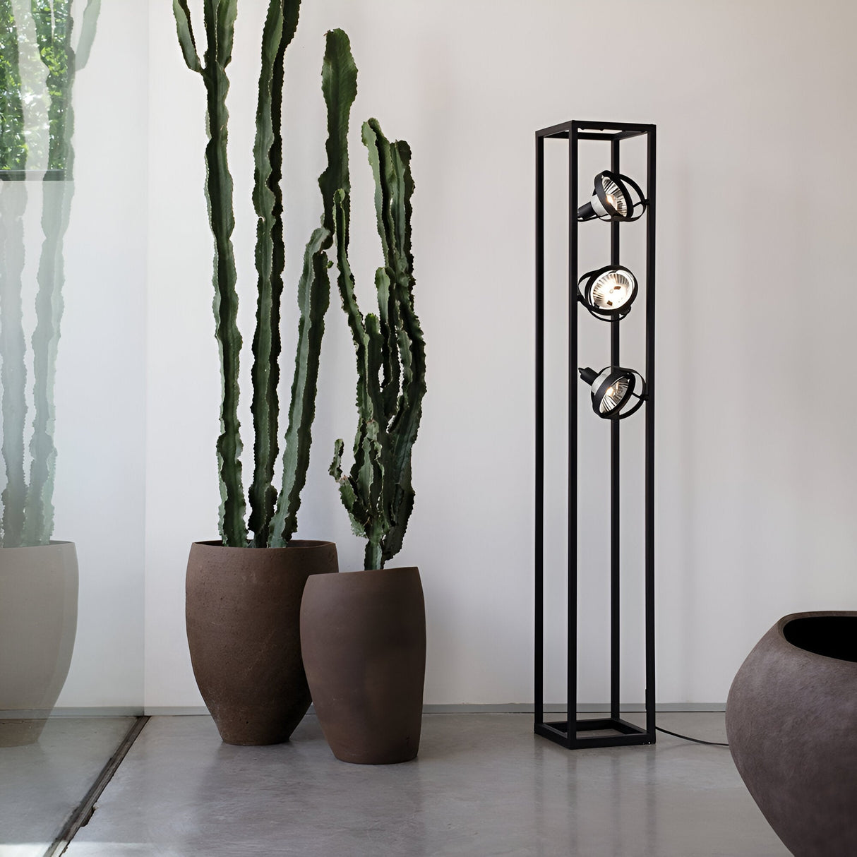 A contemporary room showcases a tall cactus in a large pot next to the elegant Zephyros Floor Lamp - Black, 130cm, which includes three spotlight bulbs set within a geometric frame. The light-toned flooring and walls are beautifully lit by natural sunlight streaming through a glass window, making it ideal for stylish modern homes.
