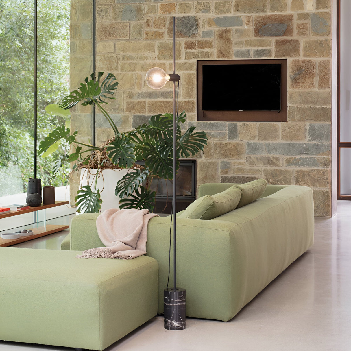 In a contemporary living room, a light green sofa is paired with the Soltus Floor Lamp in black, boasting a marble base. A potted plant enhances the minimalist decor, while a stone wall serves as the backdrop for a flat-screen TV. Expansive windows offer views of vibrant greenery outside, and a beige throw blanket is elegantly draped over the sofa.