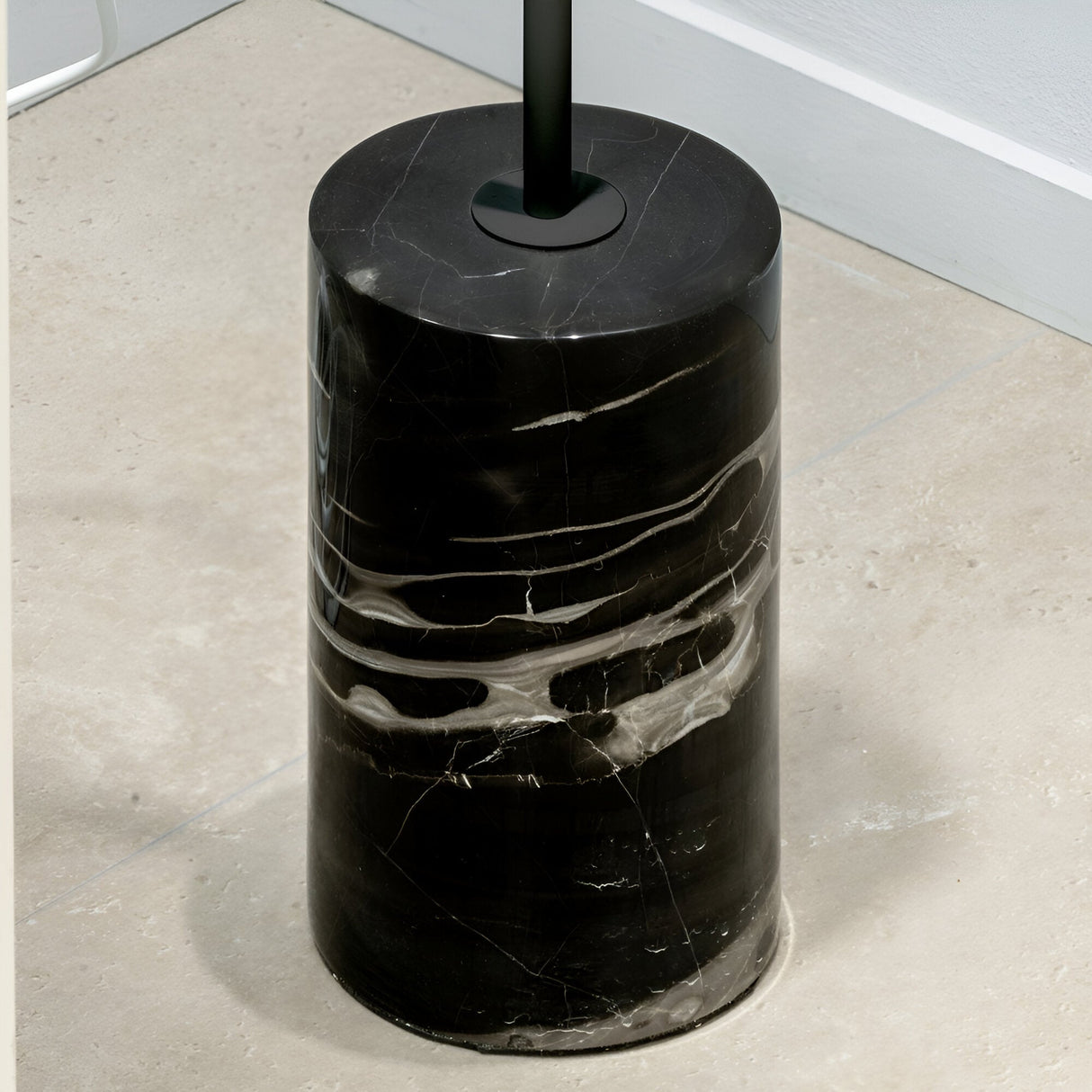 A sleek, cylindrical black marble toilet brush holder with white veining is positioned on a light beige tiled floor against a white wall. Its minimalist design mirrors the elegance of the Soltus Floor Lamp in black, showcasing a harmonious handle extending from the top.