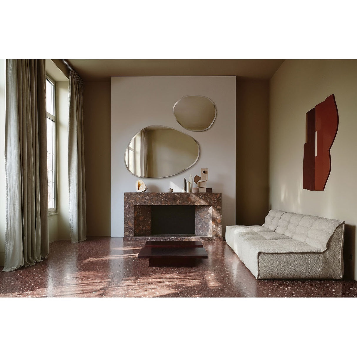 Frameless organic wall mirror with bevelled edge above a marble fireplace in a living room, styled with a second oval mirror and cream tufted sofa.