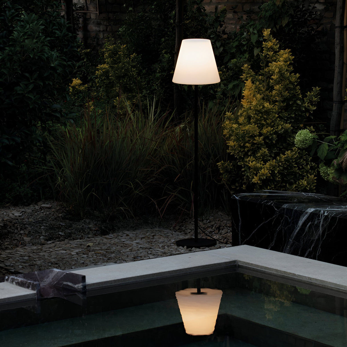 A serene garden scene at night features the Shine Outdoor Floor Lamp in grey and white, casting a soft glow over lush greenery and yellow plants. This weather-resistant lighting reflects on the surface of a nearby pond with a smooth stone edge, enhancing the tranquil atmosphere.
