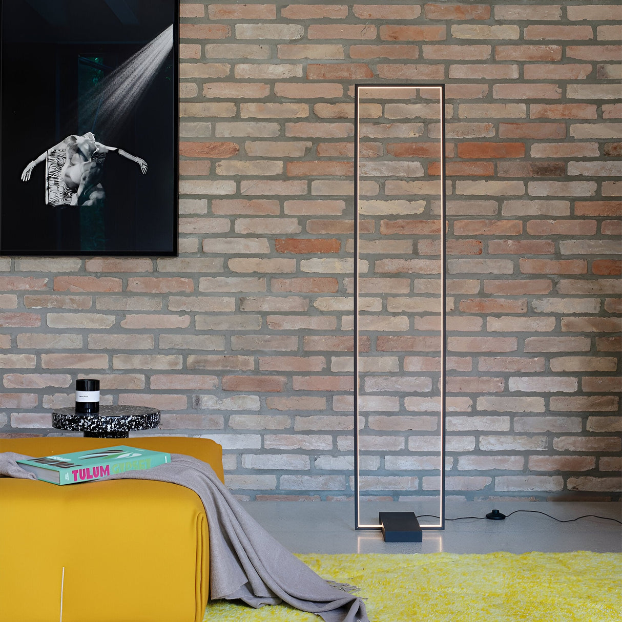 A modern living space with a brick wall showcases the Prios LED Floor Lamp 30W 3000K in black, accompanied by a black and white framed photo. The minimalist design elements include yellow cushioned seating draped with a gray blanket and a small round table decorated with books and a candle.