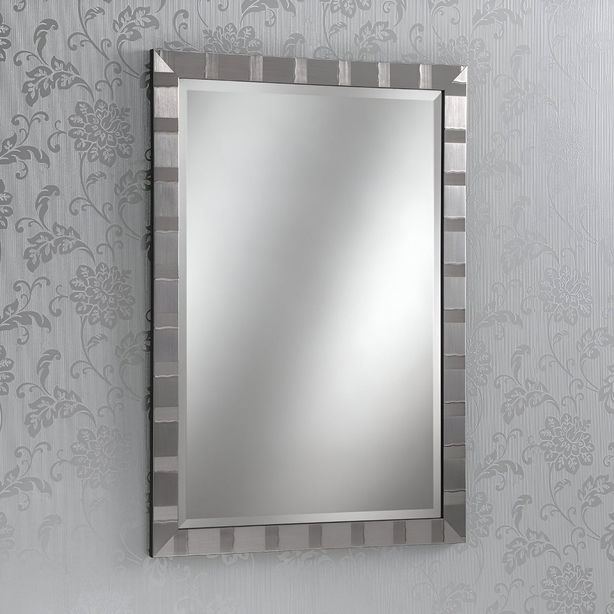 The Vault Rectangle Wall Mirror - Grey, featuring a striped grey frame, is mounted on a wall adorned with floral-patterned wallpaper. Its contemporary design beautifully reflects light, enhancing the room's bright and minimalistic look.