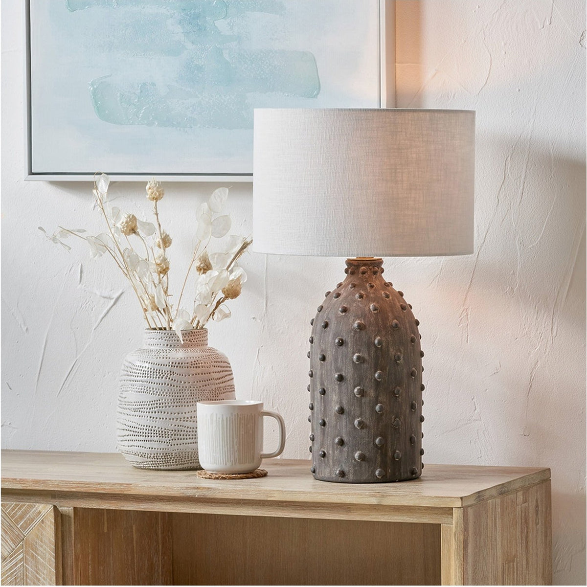 Kali Textured Dot Grey Stoneware Table Lamp Base, 37cm, with linen drum shade, styled on a wooden sideboard beside a patterned vase and mug in a living room setting.