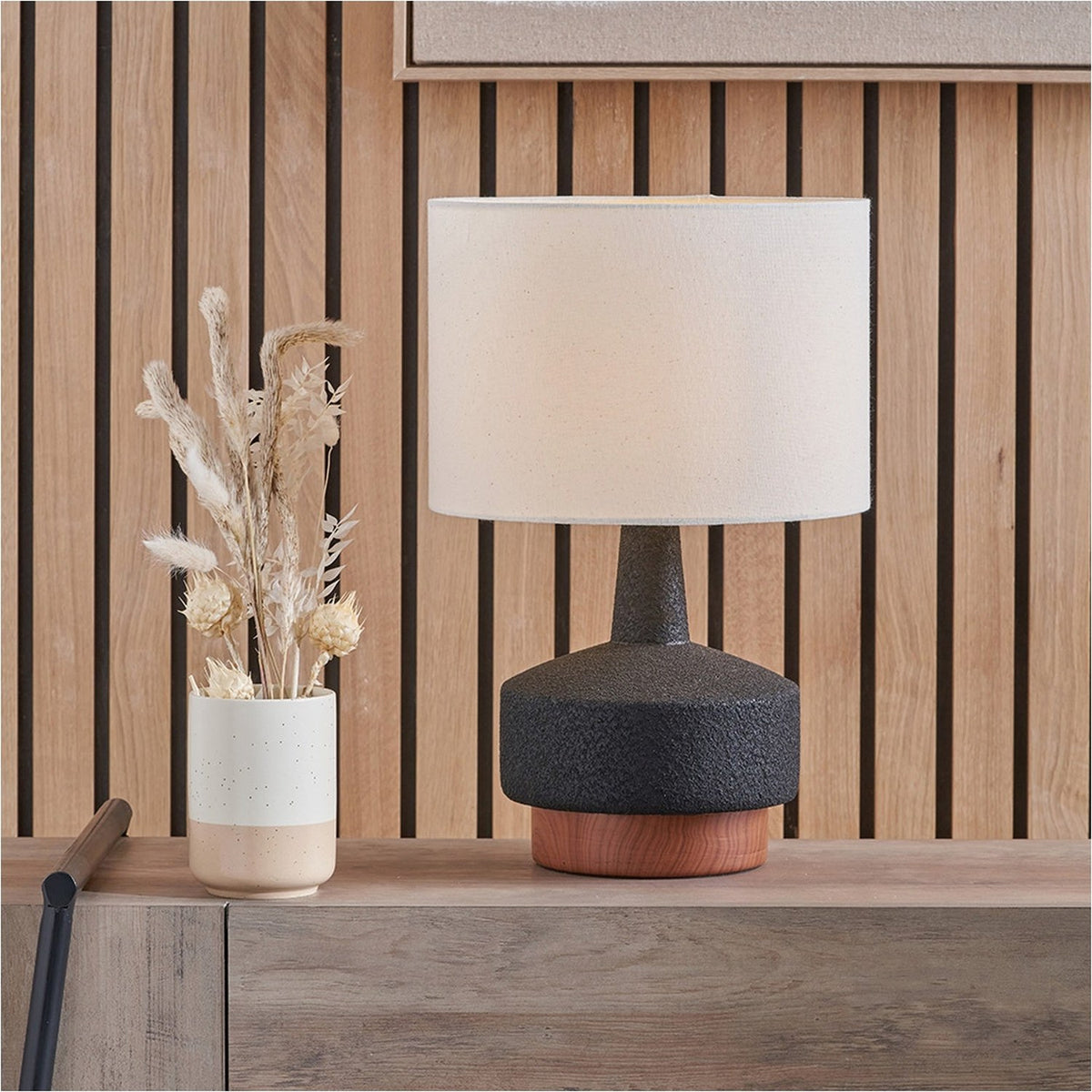 Elio Black & Wood Effect Textured Ceramic Table Lamp, 43cm, with a cream drum shade on a wood base, shown on a sideboard against a wooden slatted wall.