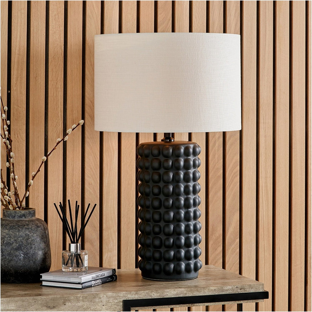 Sohan black bobbled ceramic table lamp with a cream drum shade, styled on a wooden console beside books and a vase against a slatted wood panel wall.
