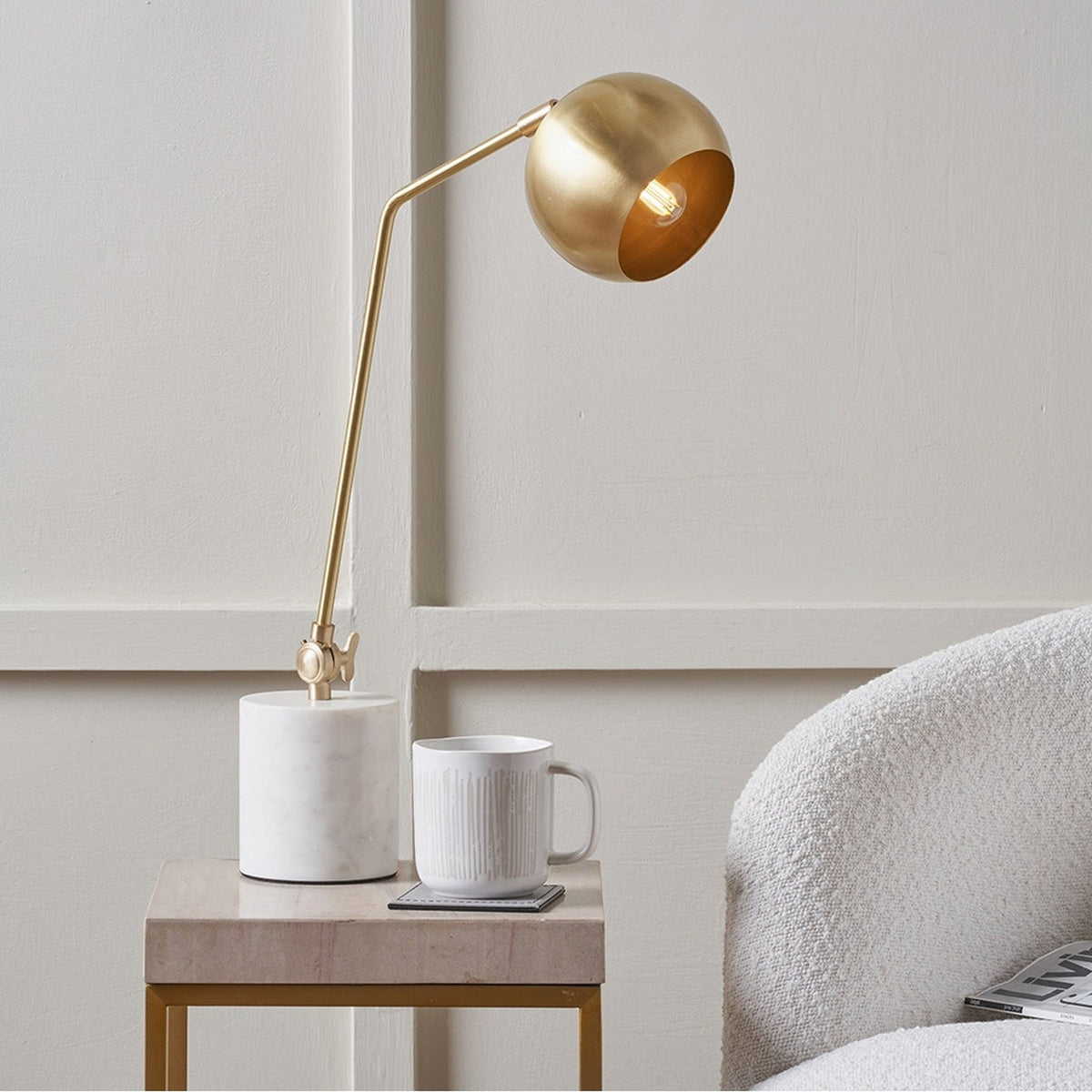 Eloise Gold Brass & Marble Table Lamp with a spherical shade on an angled arm and round marble base, shown bedside on a stone side table.