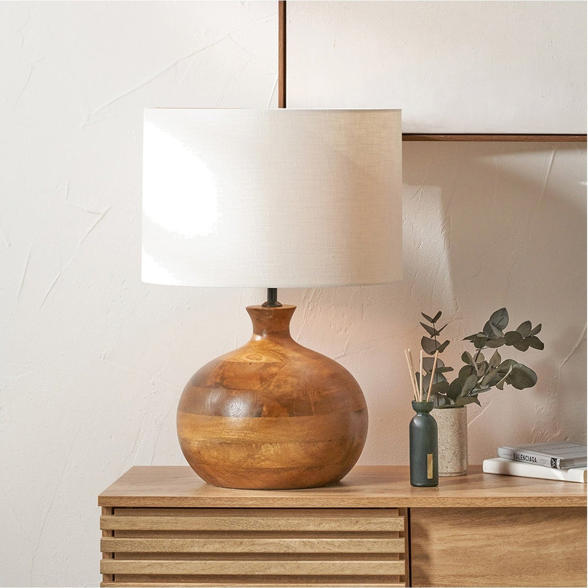 Kiah table lamp with round mango wood base in an oiled brown finish and a white drum shade, styled on a wooden sideboard.