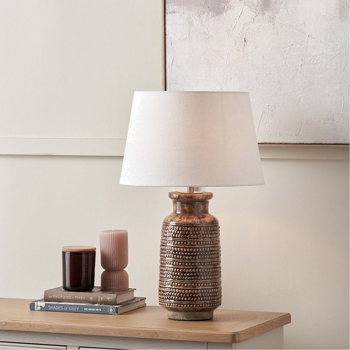 Theano Dot Design Brown Glaze Tall Table Lamp Base with textured ceramic body and white tapered shade, shown on a wooden sideboard beside books and candles.
