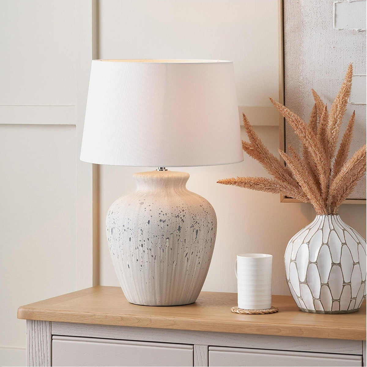 Kalli white and grey flecked ceramic table lamp, 54cm, with white fabric drum shade, styled on a wooden sideboard beside a patterned vase and dried pampas grass.