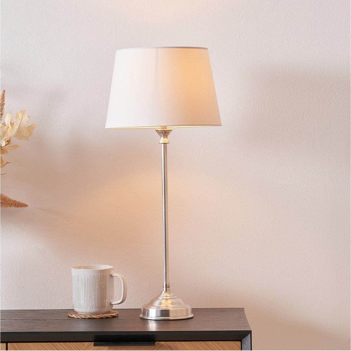 Auden Silver Metal Stick Table Lamp Base with white tapered shade, styled on a dark sideboard beside a mug in a bedroom setting.
