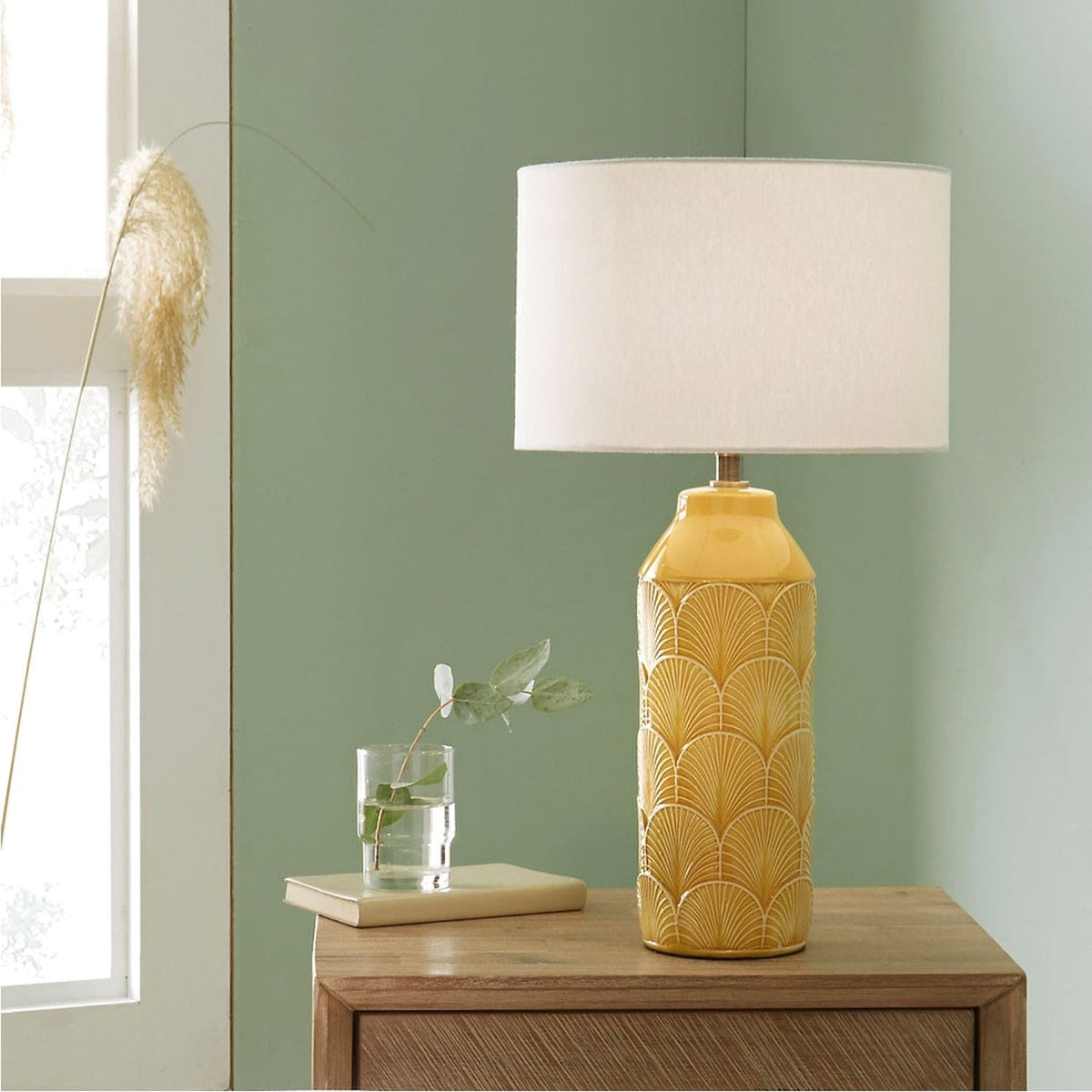 Bethan Embossed Mustard Yellow Ceramic Table Lamp, 51cm, with art deco fan pattern and white linen drum shade, shown on a wooden sideboard in a green-walled room.