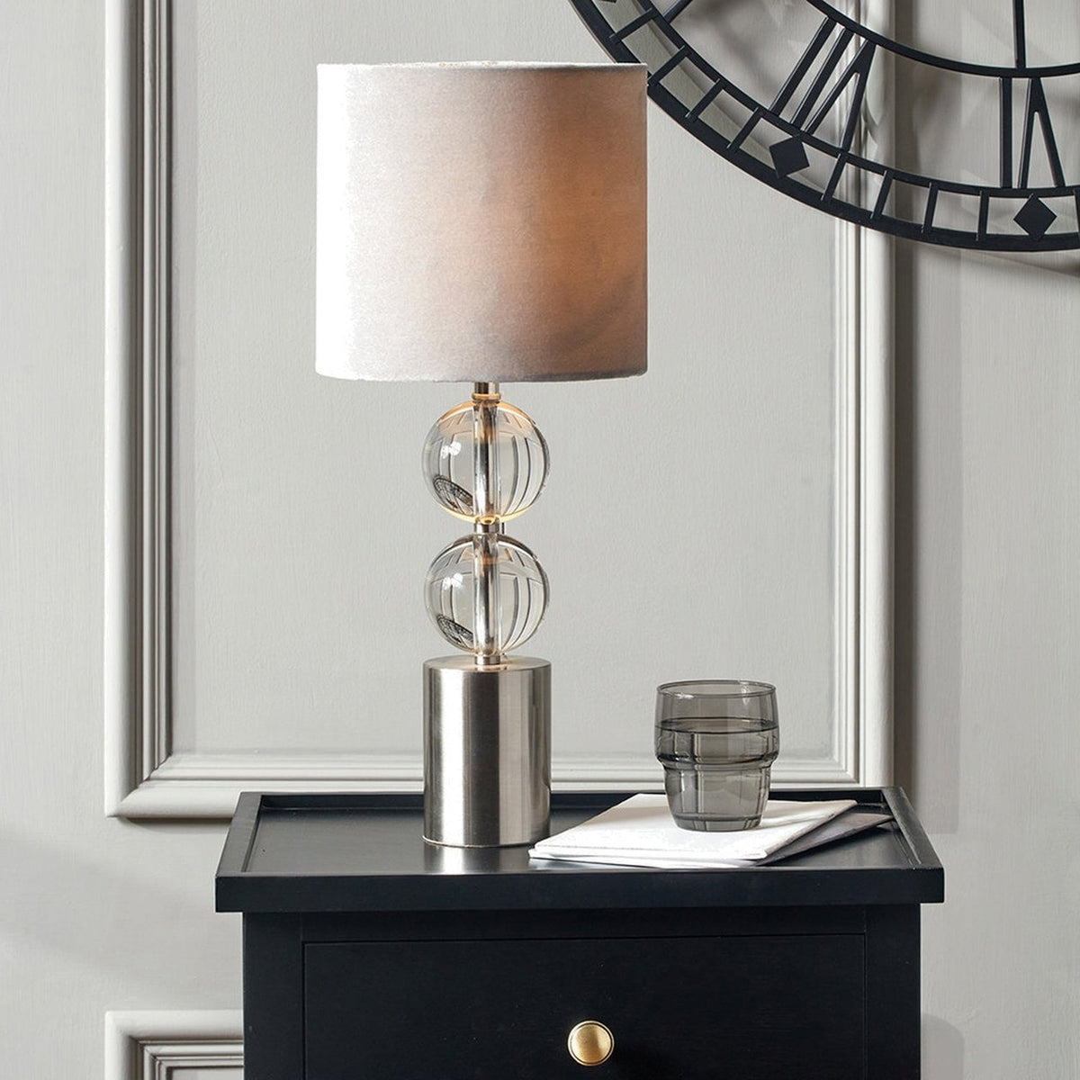 Harris Brushed Silver & Clear Glass Table Lamp, 49cm, with stacked glass sphere stem and grey drum shade, shown on a black bedside cabinet beneath a wall clock.