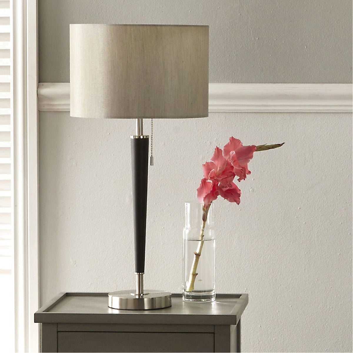 Lowry table lamp with brushed silver base and matt black iron stem, oval fabric shade, shown on a nightstand beside a glass vase with pink flowers.