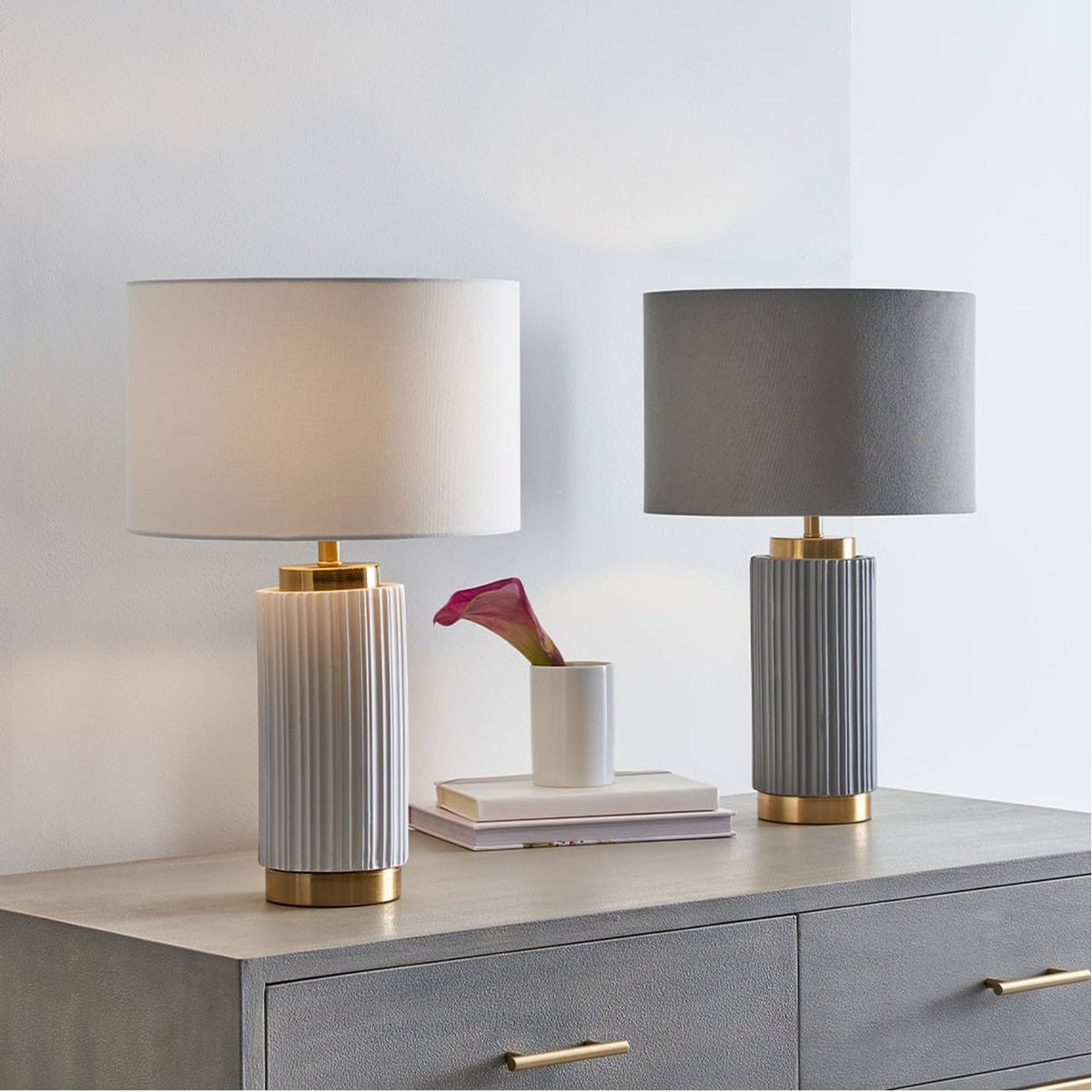 Ionic Grey Textured Ceramic & Gold Metal Table Lamp with a grey drum shade, shown on a chest of drawers beside a matching white lamp in a bedroom setting.