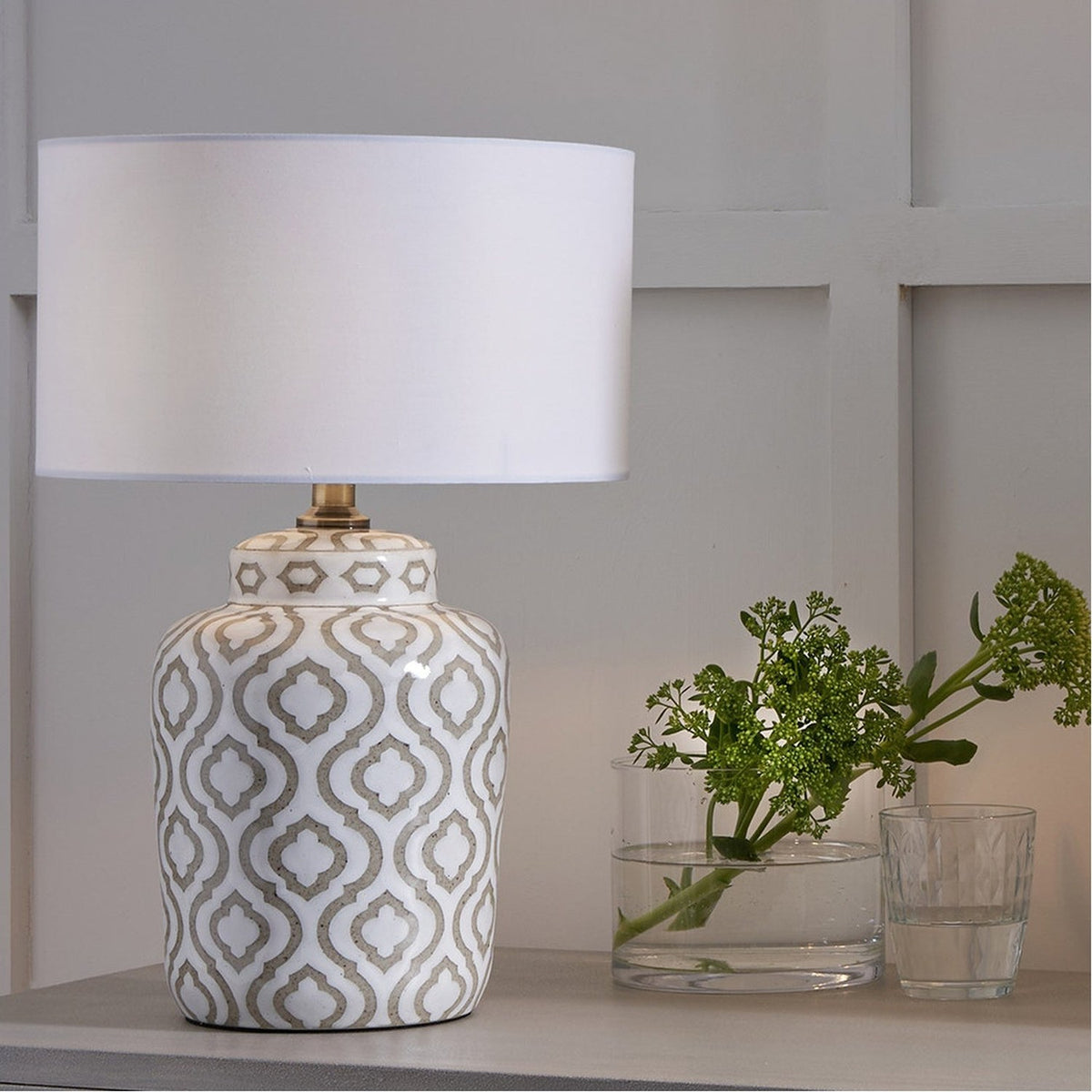 Celia Taupe & White Pattern Ceramic Table Lamp with white drum shade, shown on a shelf beside a plant in a vase in a neutral-toned room.
