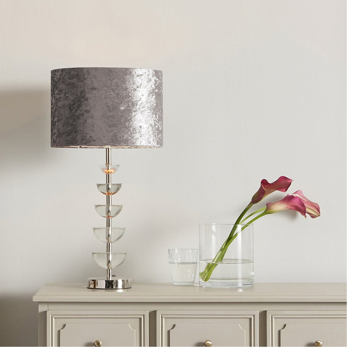 Rosa Glass & Silver Table Lamp Base, 45cm, with stacked clear glass discs on a silver metal stem and grey shade, styled on a cabinet beside a vase and calla lilies.