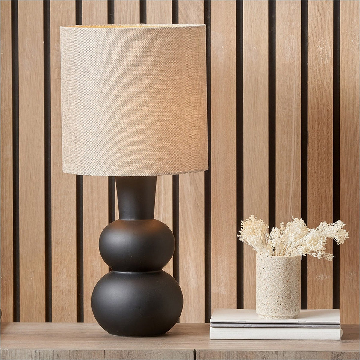 Aaliyah Black Curved Bottle Ceramic Table Lamp, 51cm, with a linen drum shade, styled on a sideboard against a slatted wood panel background.