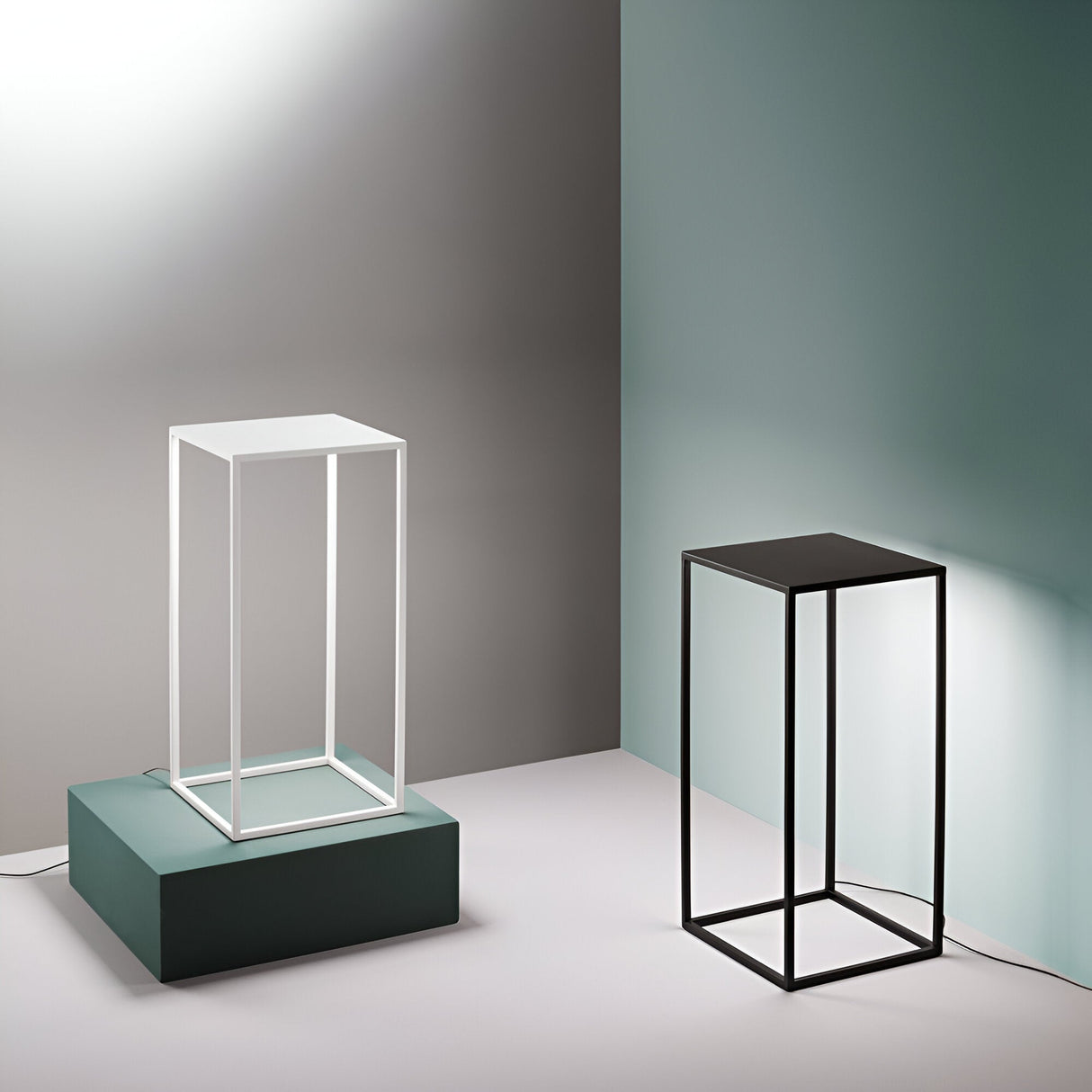 In a minimalist interior scene, two cube-shaped frames are displayed on the floor. The white frame is positioned atop a green podium, while the black frame rests directly on the ground. The sophisticated setting is enhanced by the 50cm Raya LED Outdoor Floor Lamp in black, with walls behind that are elegantly two-toned in grey and teal.