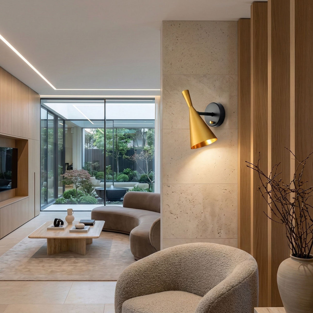Modern living room with neutral tones, curved sofa, cozy armchair, wood coffee table, built-in shelves, and glass doors to a lush garden. The Funk Wall Light - Gold brings luxury lighting to the stone accent wall.
