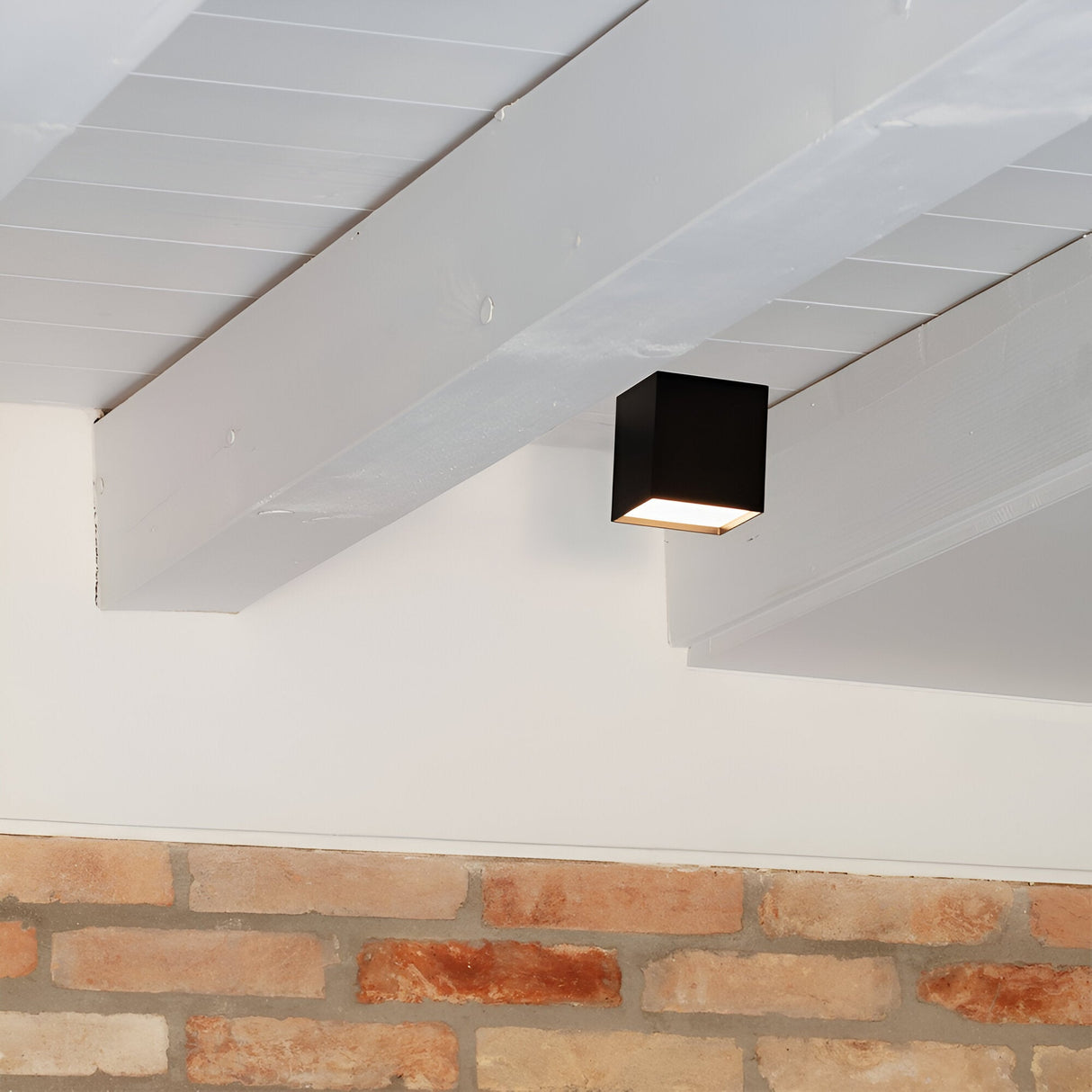 A Luminex LED Downlight 10W 3000K in black, featuring a minimalist square design, is mounted on white wooden beams. Below, an exposed brick wall provides contrast to the sleek ceiling design. The setting evokes a contemporary interior space with energy-efficient performance.
