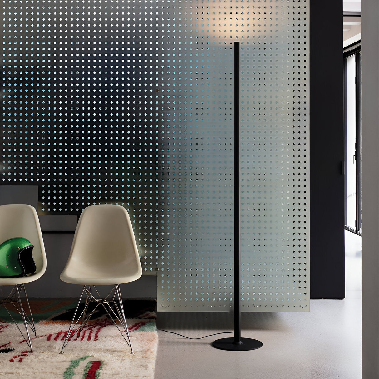 A modern interior showcases the Noctis LED Floor Lamp 24W 3000K - Black, with its adjustable lighting, placed next to a perforated panel. Two white chairs and a green helmet sit on a patterned rug, contributing to the sleek, contemporary design.