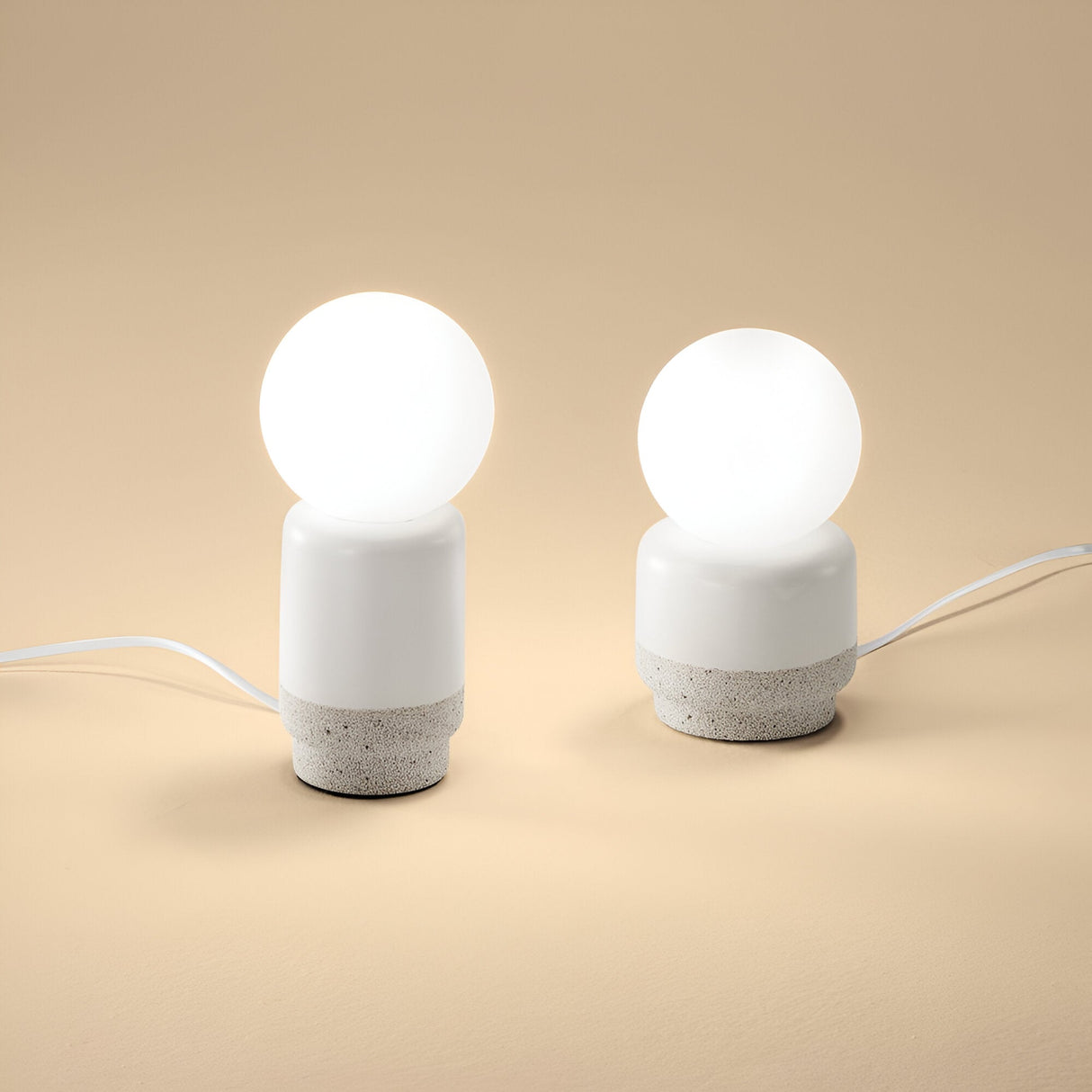 Two Brillara Small Table Lamps with spherical white bulbs and cylindrical cement bases rest on a smooth beige surface. These industrial-style lamps, connected by white cords, stand out as chic accent lighting against the neutral backdrop.